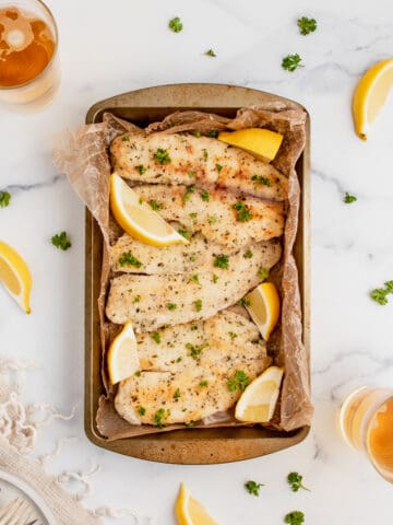 Pan fried fish in a baking dish with fresh herbs and lemon wedges.