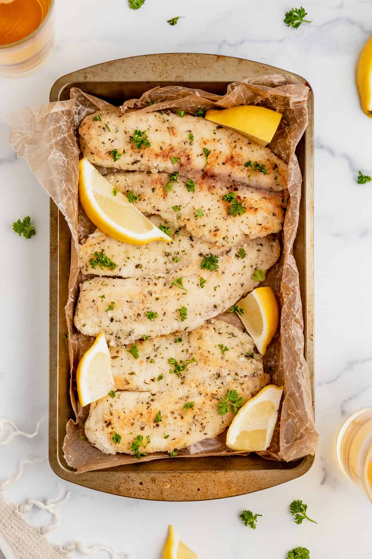 Pan fried fish in a baking dish with fresh herbs and lemon wedges.