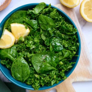 Blue bowl filled with a spinach and kale salad, with lemon wedges on top.