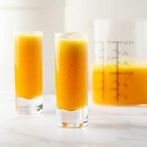 A featured image of two ginger turmeric shots with a measuring cup of the extra juice in the back.