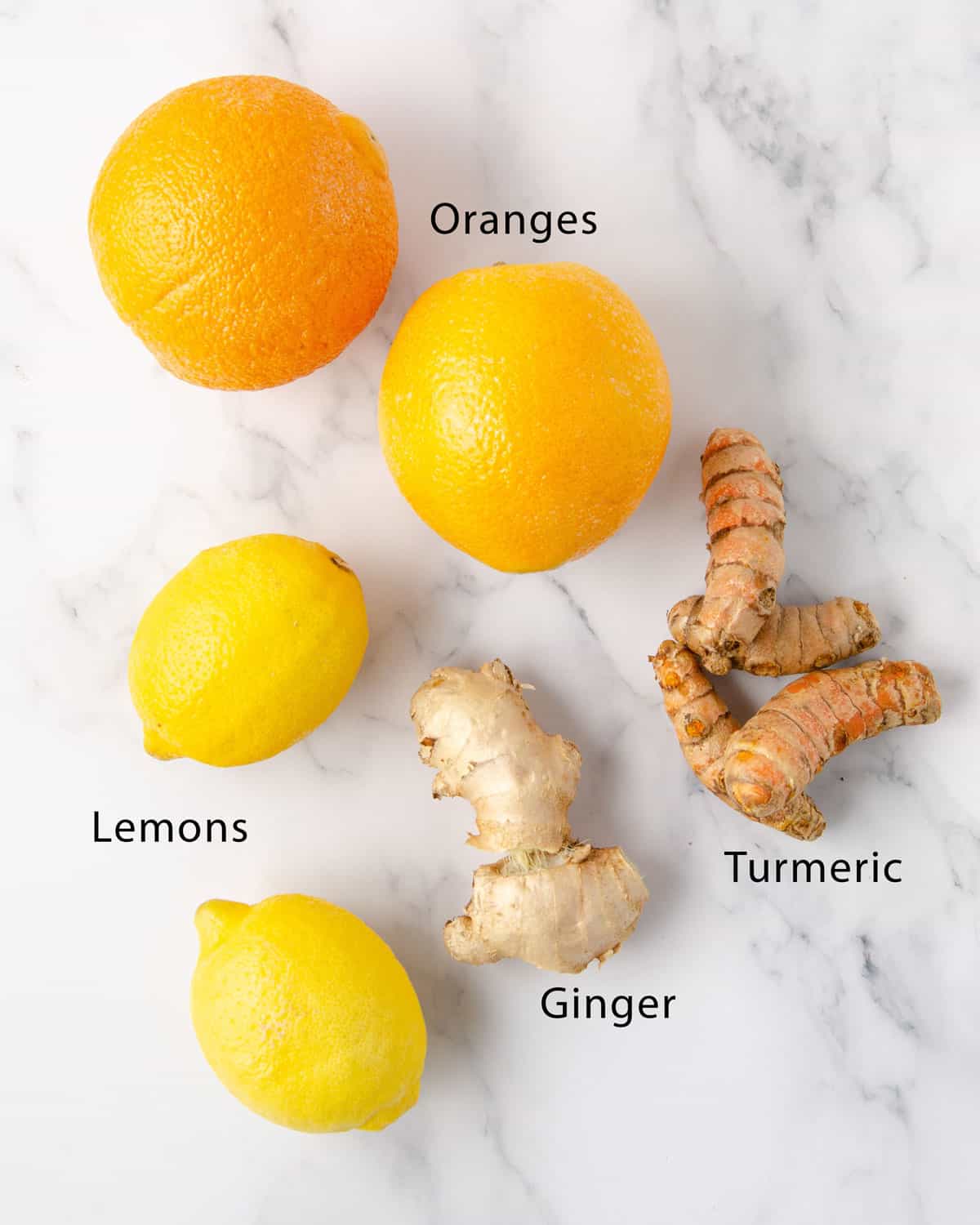 Two lemons, two oranges, 4 pieces of turmeric and two pieces of ginger on a marble countertop to represent the ingredients needed for a ginger turmeric shot.