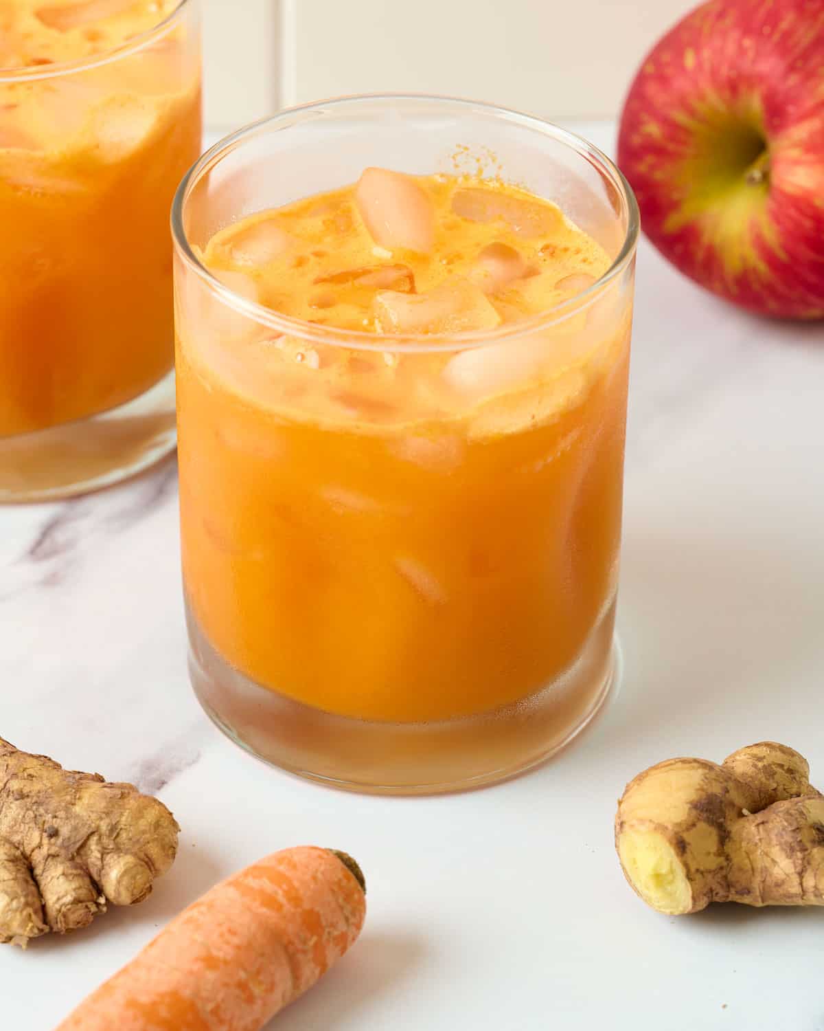 A cold glass of carrot ginger turmeric juice with carrots, ginger, and an apple around it.