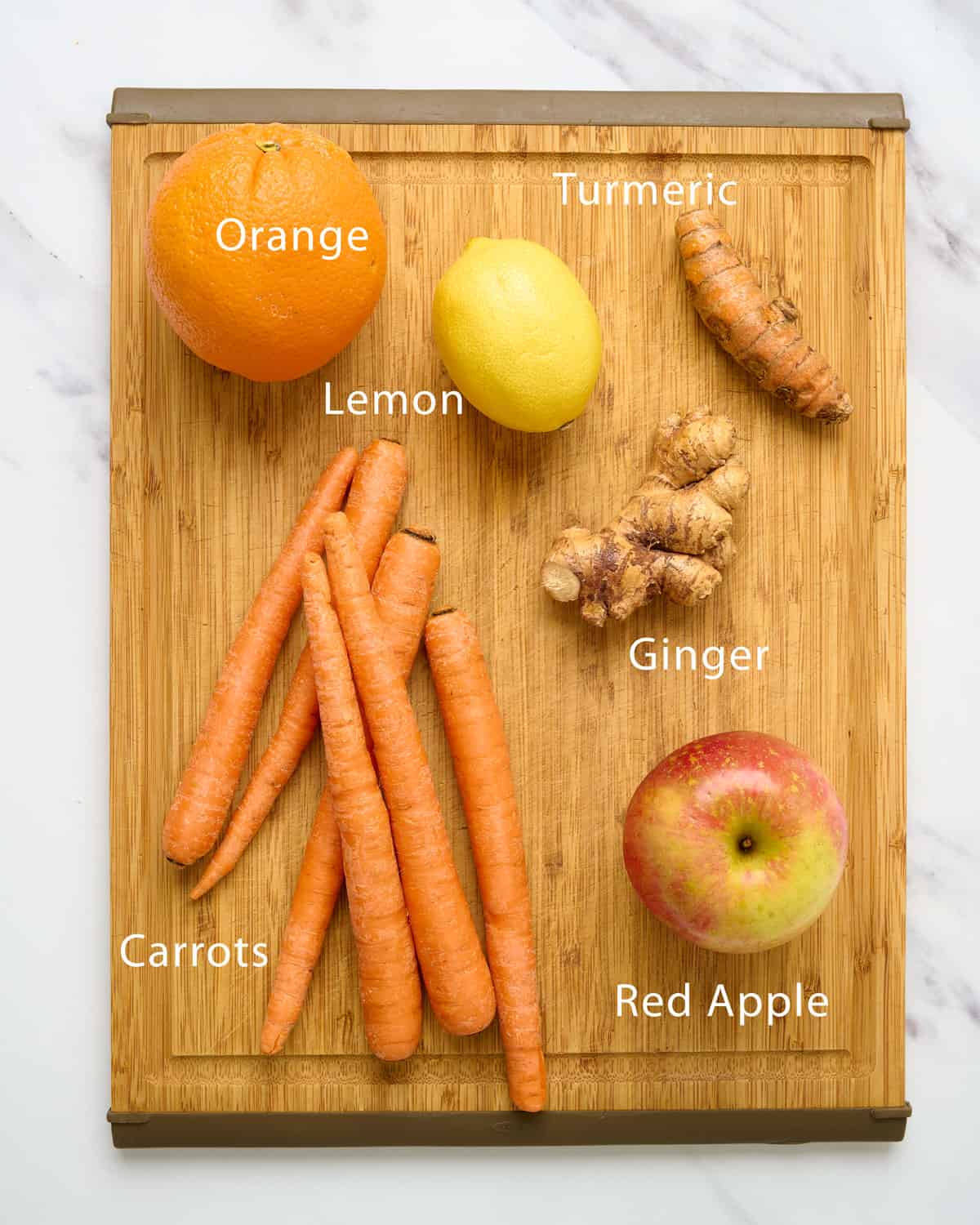 An orange, lemon, carrots, ginger, turmeric, and an apple on a cutting board.